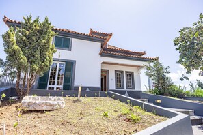 Ein zweistöckiges Gebäude mit traditionellem Ziegeldach und grünen Fensterläden, umgeben von einem angelegten Garten mit verschiedenen Pflanzen und einem Kiesweg.