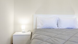 Una cama bien hecha con colcha gris, una mesita de noche blanca con una lámpara y una pared blanca.