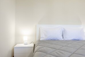 Ein ordentlich bezogenes Bett mit einer grauen Decke, ein weißer Nachttisch mit einer Lampe und eine weiße Wand.