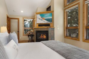 A bedroom with a fireplace, a bed with pillows, a table with framed photos, and a painting of a skier. - Elevated Eagle-Vail Duplex with Hot Tub (Avon)
