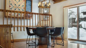 Uma sala de jantar com mesa e cadeiras de madeira, um lustre e vista para uma paisagem nevada através de uma porta de vidro.