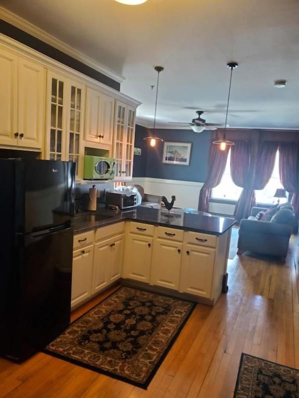 A kitchen with white cabinets, a black refrigerator, and a microwave. There is a rug on the floor and a ceiling fan.