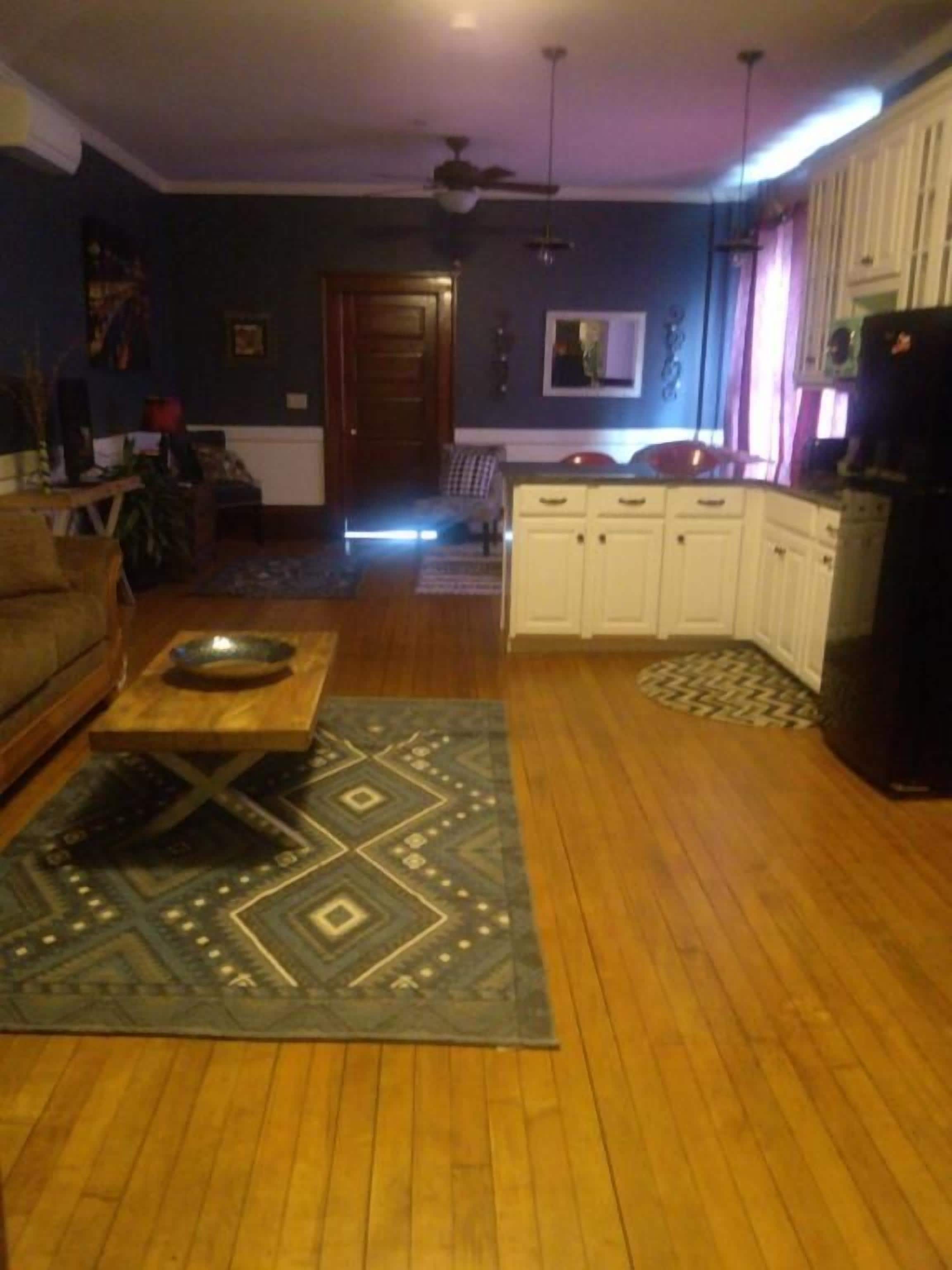 A living room with a wooden floor, a dining table with a patterned rug, and a kitchen area with white cabinets and a refrigerator.