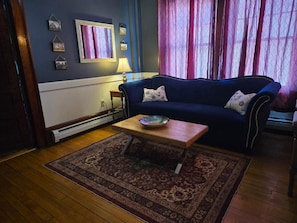 A living room with a dark blue sofa, a wooden coffee table, a patterned rug, and a window with pink curtains.