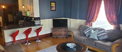 Un salon avec un canapé, une table basse et un téléviseur. On aperçoit des fauteuils rouges et un meuble en arrière-plan.