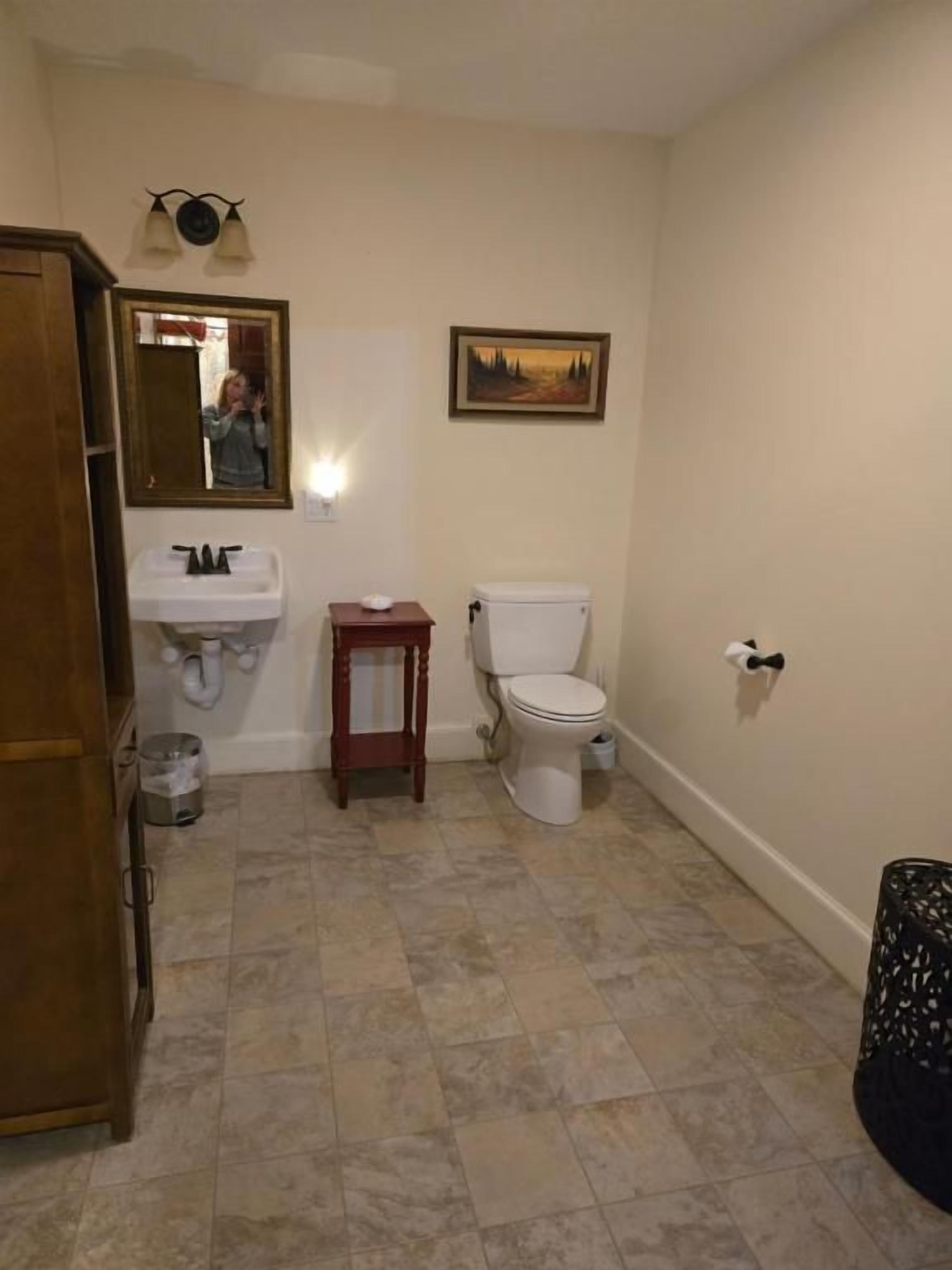 Un baño con inodoro, lavamanos, espejo y una pequeña mesa.