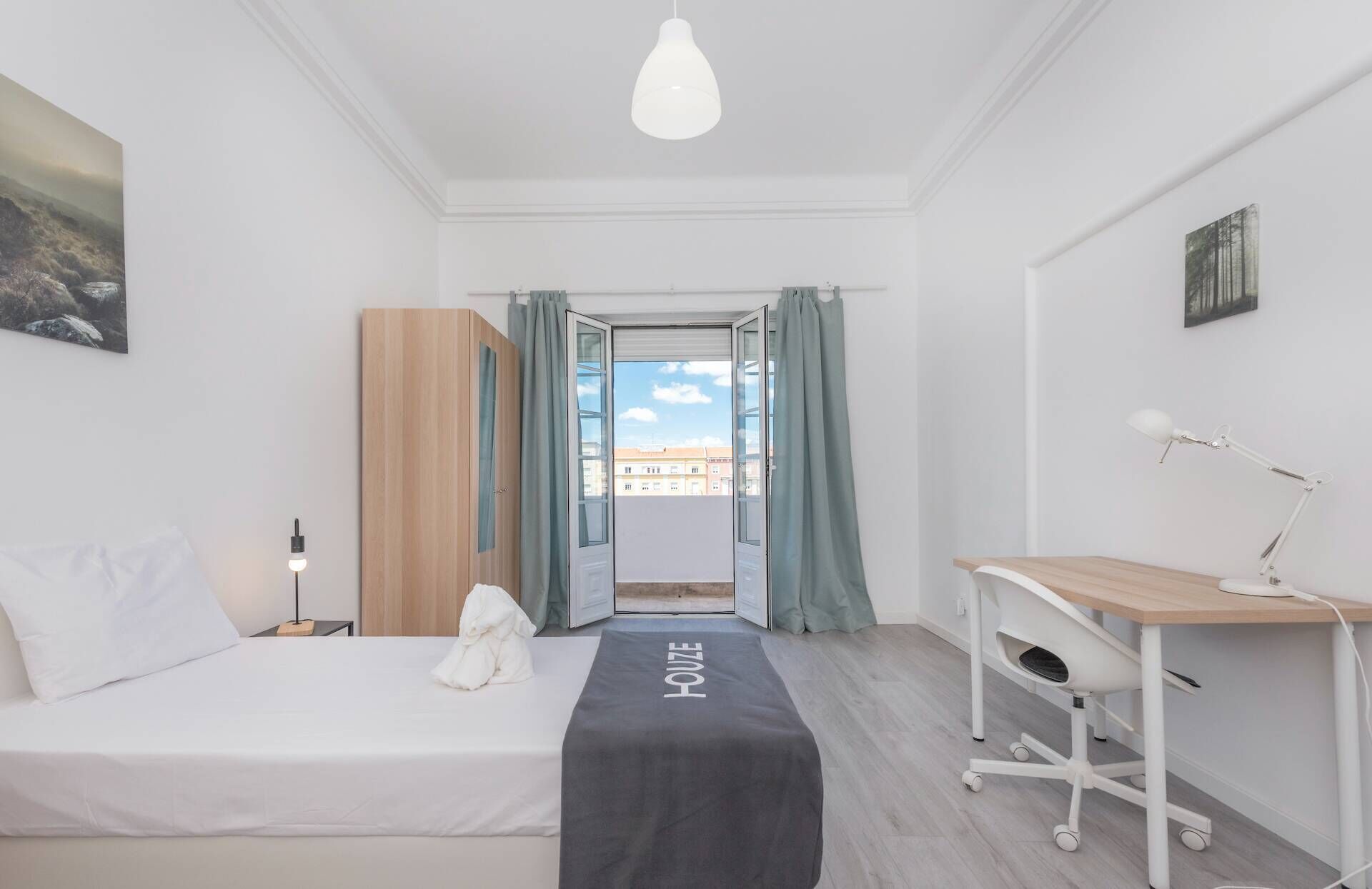 Ein Hotelzimmer mit einem Bett, einem Schreibtisch, einem Stuhl, einer Lampe und Blick durch eine offene Tür.