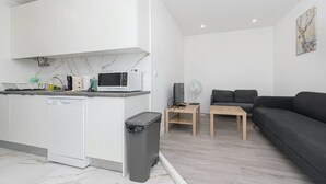 Una cucina moderna con pensili bianchi, un forno a microonde e una lavastoviglie. Nell'area soggiorno, si trova un piccolo tavolo con una ventola e un divano grigio.