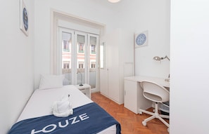 A hotel room with a bed, a desk, a chair, a mirror, and a window with a view of buildings. - HOUZE Apartment in Historical Lisbon (Lisboa)