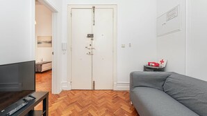 Ein Zimmer mit Fernseher auf einem Schrank, einem grauen Sofa, einem Holzboden mit Fischgrätmuster und einer weißen Tür mit Schloss.