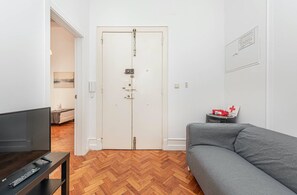 A room with a television on a stand, a grey sofa, a wooden herringbone floor, and a white door with a lock. - HOUZE Apartment in Historical Lisbon (Lisboa)