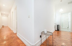 Um corredor com piso de madeira, paredes brancas e uma pequena mesa com uma xícara.