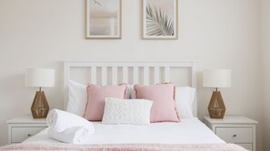 Un letto rifatto con lenzuola bianche e rosa, affiancato da due comodini con lampade e cesti.