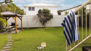 Un giardino sul retro con una casa bianca, un pergolato in legno, un piccolo albero e un asciugamano a righe su una ringhiera.