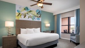 Ein Hotelzimmer mit einem Bett, zwei Nachttischen, einem Deckenventilator, einem großen Fenster mit Vorhängen und einem Sessel.