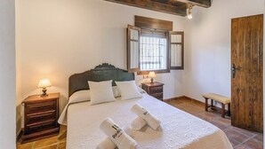 A neatly made bed with white linens and two pillows, a wooden headboard, a nightstand with a lamp, a bench, and a wooden door. - Huerta San Francisco Casarabonela by Ruralidays (Málaga)