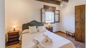 Ein ordentlich bezogenes Bett mit weißen Leinen und zwei Kissen, ein Holz-Kopfteil, ein Nachttisch mit Lampe, eine Bank und eine Holztür.