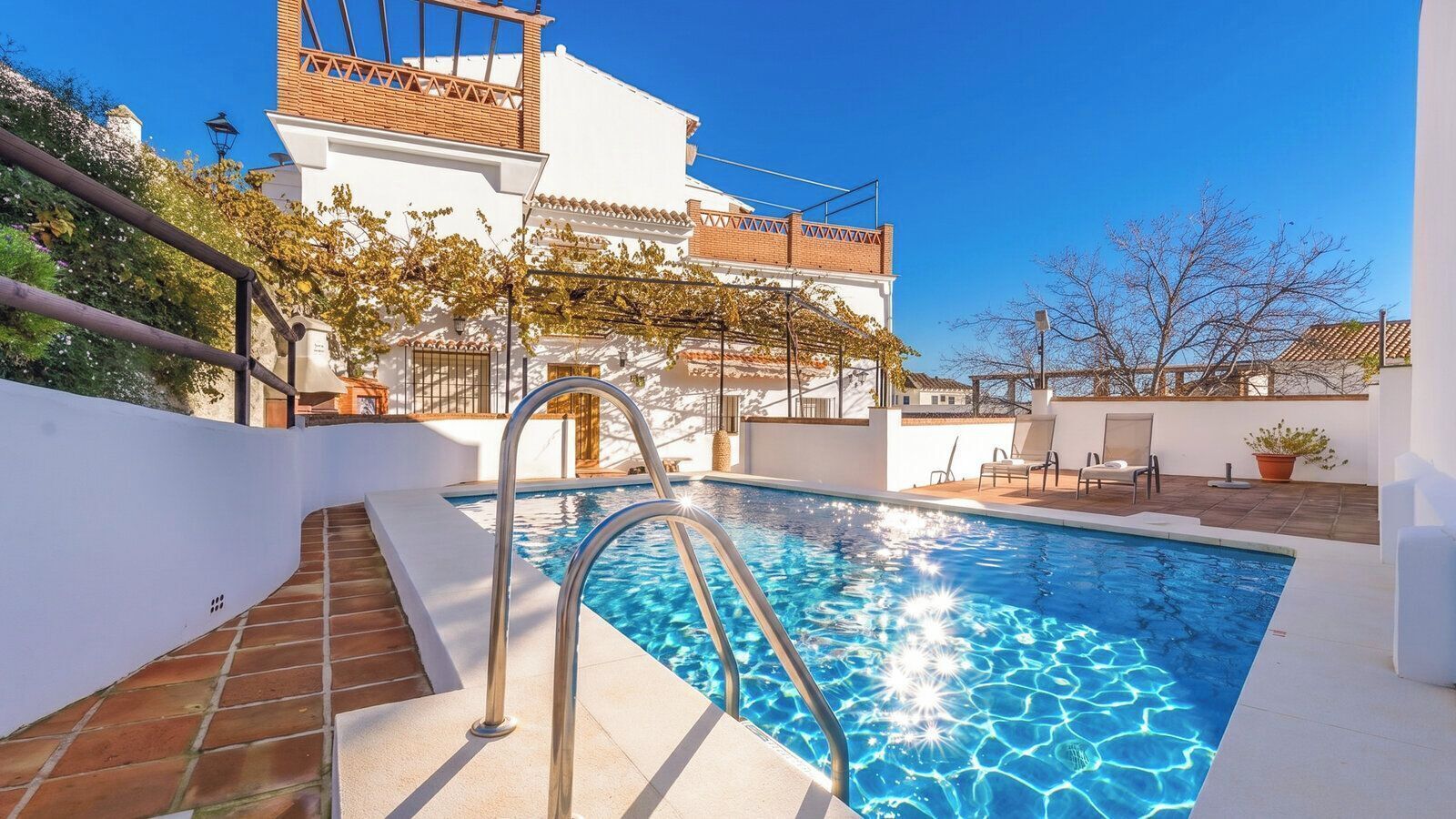 Une piscine avec un escalier en métal, un bâtiment blanc avec un toit recouvert de tuiles et un ciel bleu clair.
