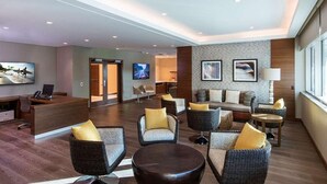 Een moderne lobby met een receptiebalie, een zithoek met rieten stoelen en banken, een televisie, kunstwerken aan de muur, en een groot raam.