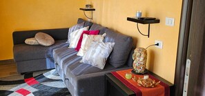 Ein Wohnzimmer mit einer Couch, Kissen, einem kleinen Tisch mit Vase und Snacks sowie einem an der Wand befestigten Regal.