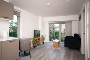 Ein modernes Wohnzimmer mit einer Couch, einem Couchtisch und einem Fernseher. Ein Fenster bietet Blick auf Grünfläche.
