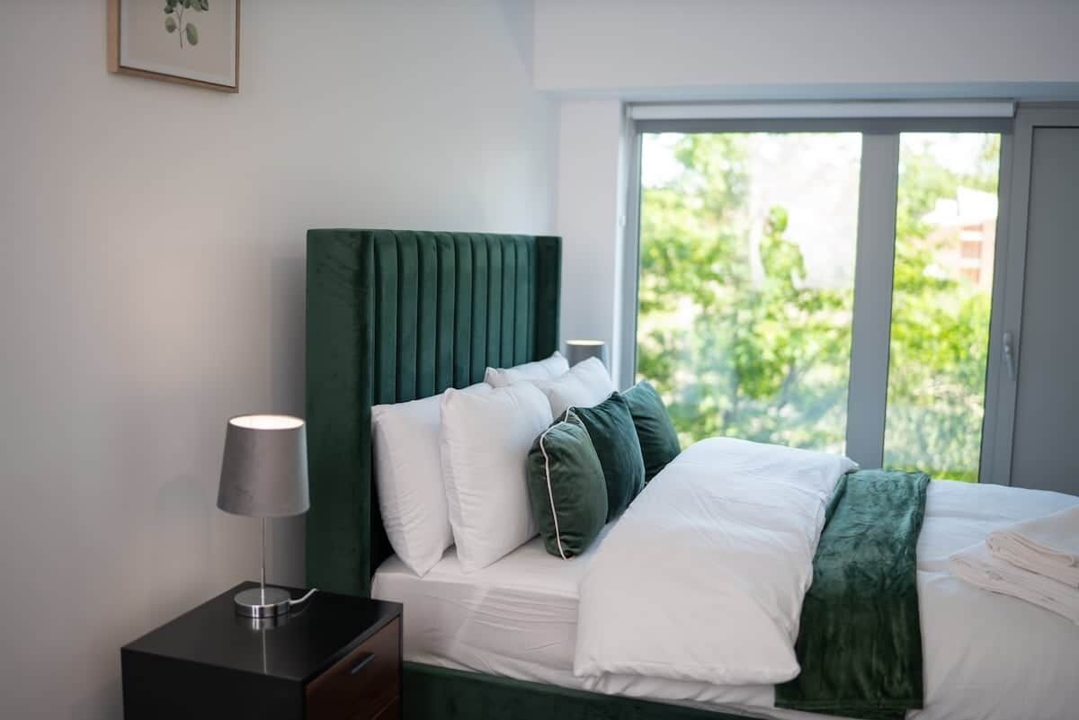 A neatly made bed with white linens and a green headboard, a bedside lamp, and a window with a view of greenery.
