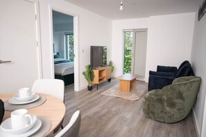 A modern room with a dining table, chairs, a sofa, a television, and a bed in the background. - Modern 2BR apartment in Manchester (Eccles)