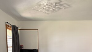 Et spiseområde med et rektangulært bord, fire stole, en loftlampe, et vindue med gardiner og et spejl monteret på væggen.