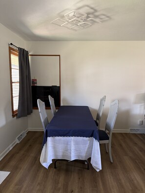 Ein Essbereich mit einem rechteckigen Tisch, vier Stühlen, einer Deckenleuchte, einem Fenster mit Vorhängen und einem an der Wand befestigten Spiegel.