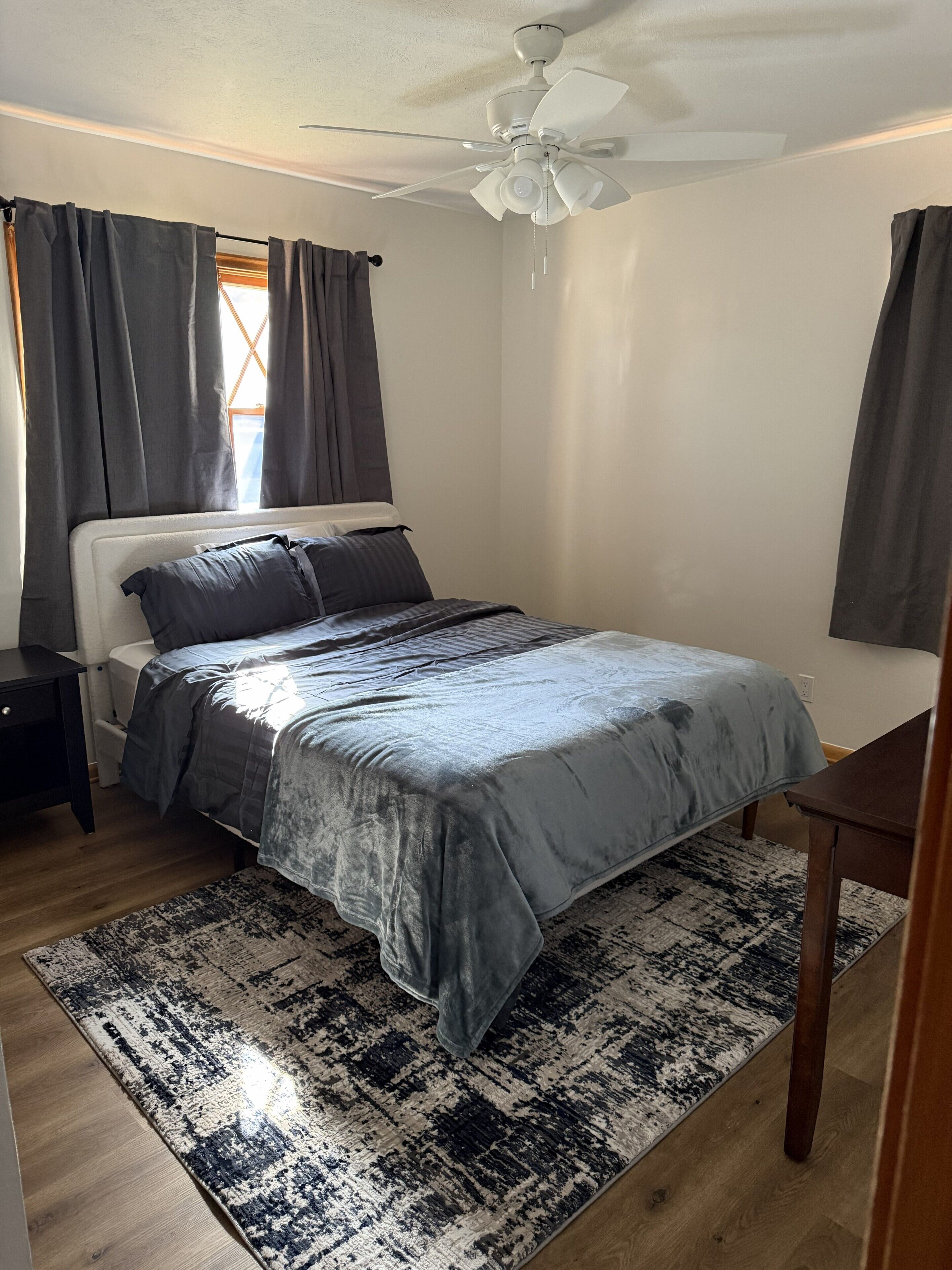 Un dormitorio con cama, mesita de noche, escritorio, ventilador de techo y ventana con cortinas.
