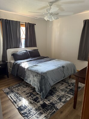 Ein Schlafzimmer mit einem Bett, einem Nachttisch, einem Schreibtisch, einem Deckenventilator und einem Fenster mit Vorhängen.