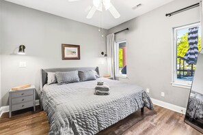 Ein Schlafzimmer mit einem Bett, Nachttischen, einem Deckenventilator, einem Fenster mit Vorhängen und einem gerahmten Bild an der Wand.