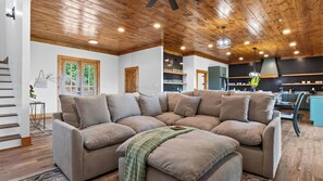 Ein modernes Wohnzimmer mit Ecksofa, Holzbalkendecke und sichtbarer Küchenzeile im Hintergrund.