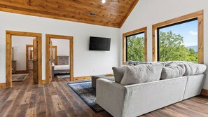 Ein modernes Wohnzimmer mit grauem Sofa, Holzboden und großen Fenstern mit Blick auf Bäume.