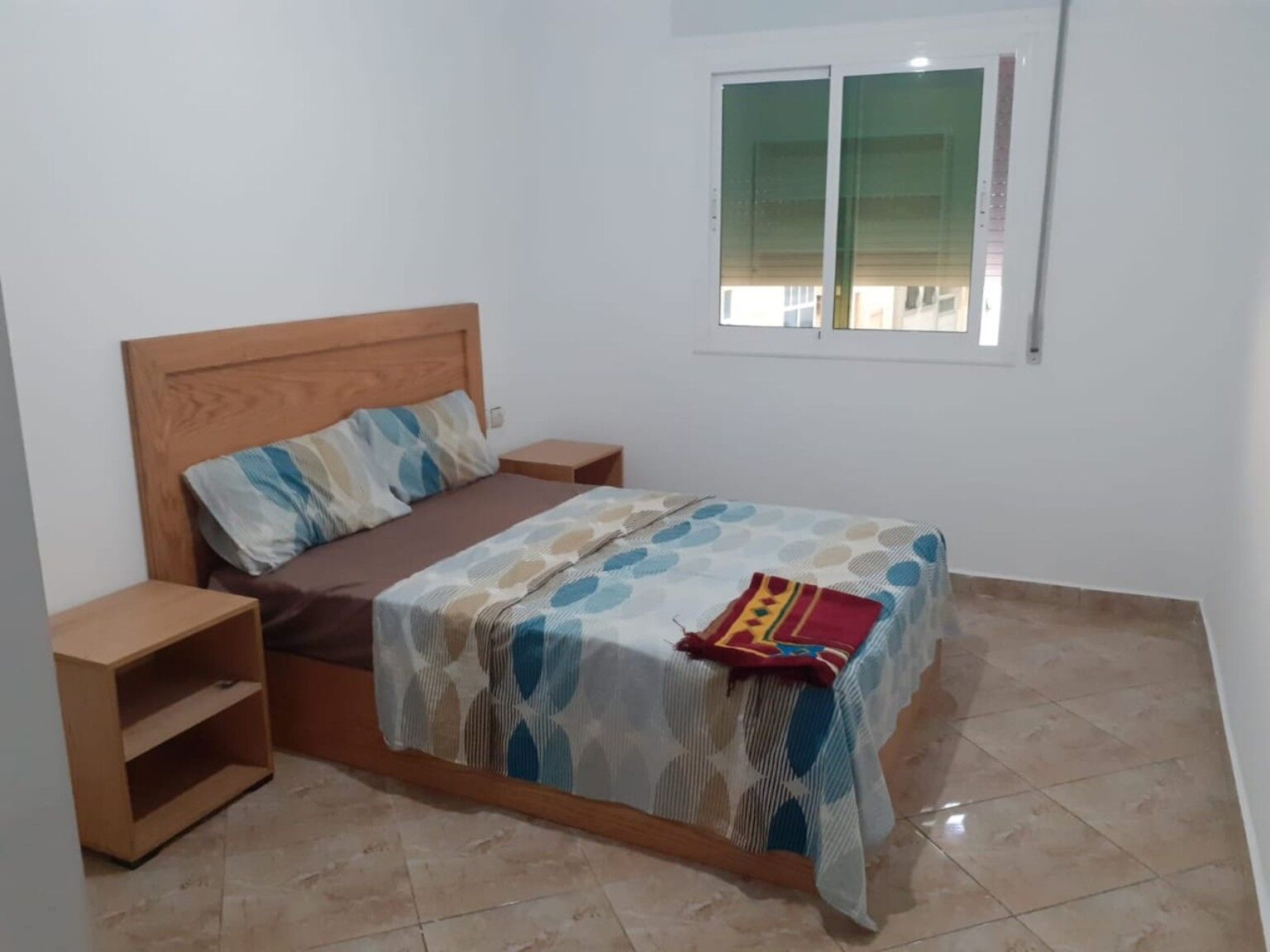 A bedroom with a wooden bed, a nightstand, a small shelf, and a window with blinds.