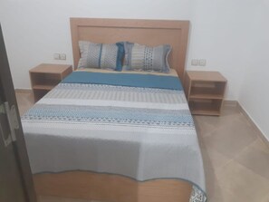 A bedroom with a wooden headboard, a bed with patterned bedding, two nightstands, and a small wooden shelf.