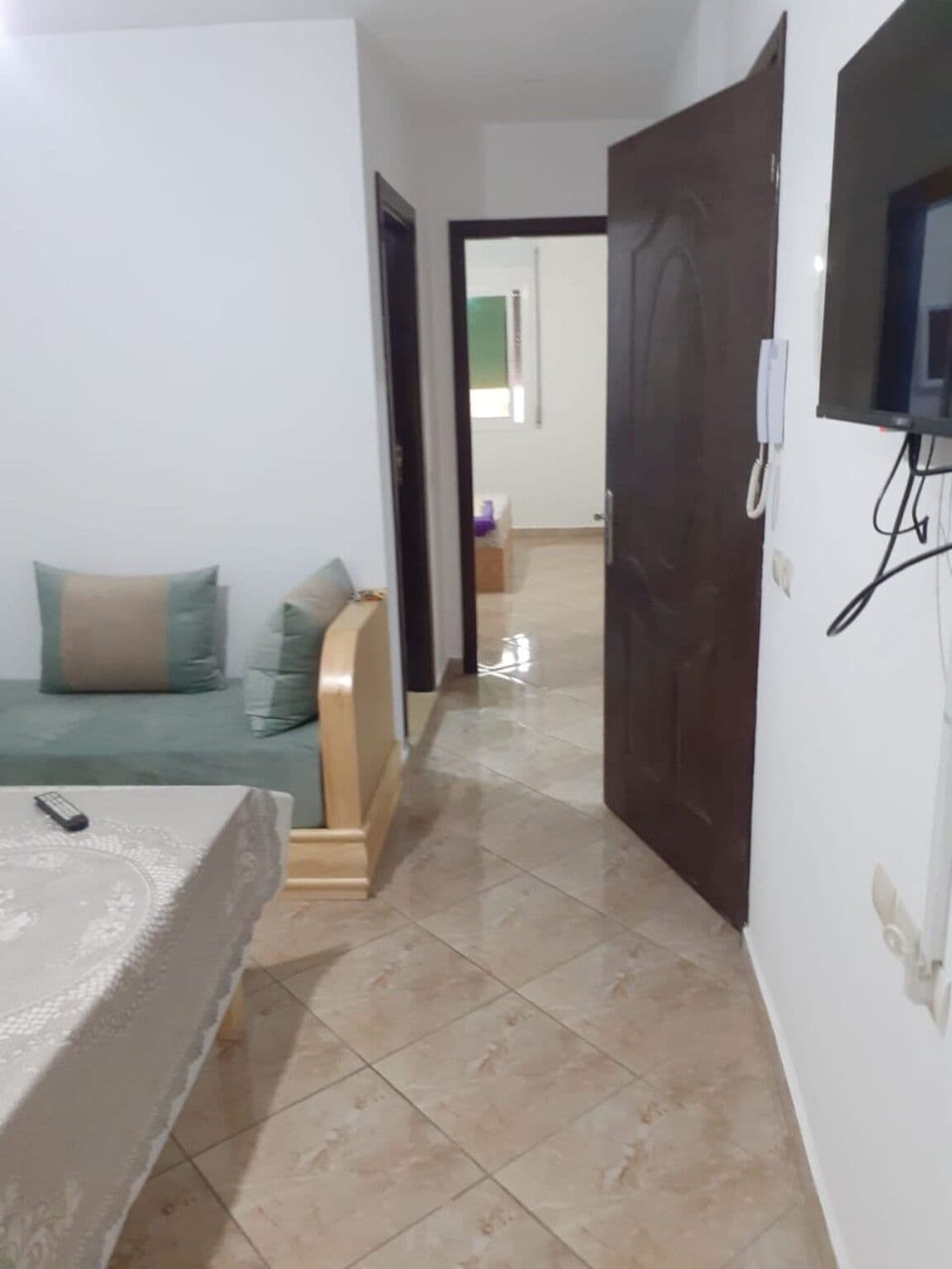 A bedroom with a bed, a bedside table, a TV mounted on the wall, and a door leading to another room.