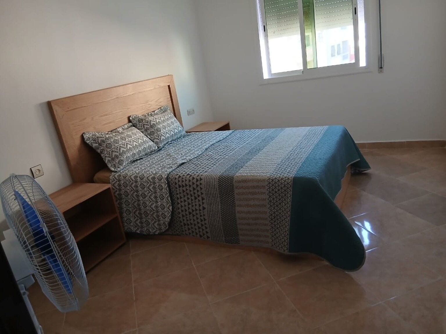 Une chambre à coucher avec une tête de lit en bois, une literie à motifs, un ventilateur sur pied et une fenêtre avec des stores.