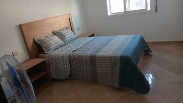 Un dormitorio con cabecera de madera, ropa de cama estampada, un ventilador de pie y una ventana con persianas.