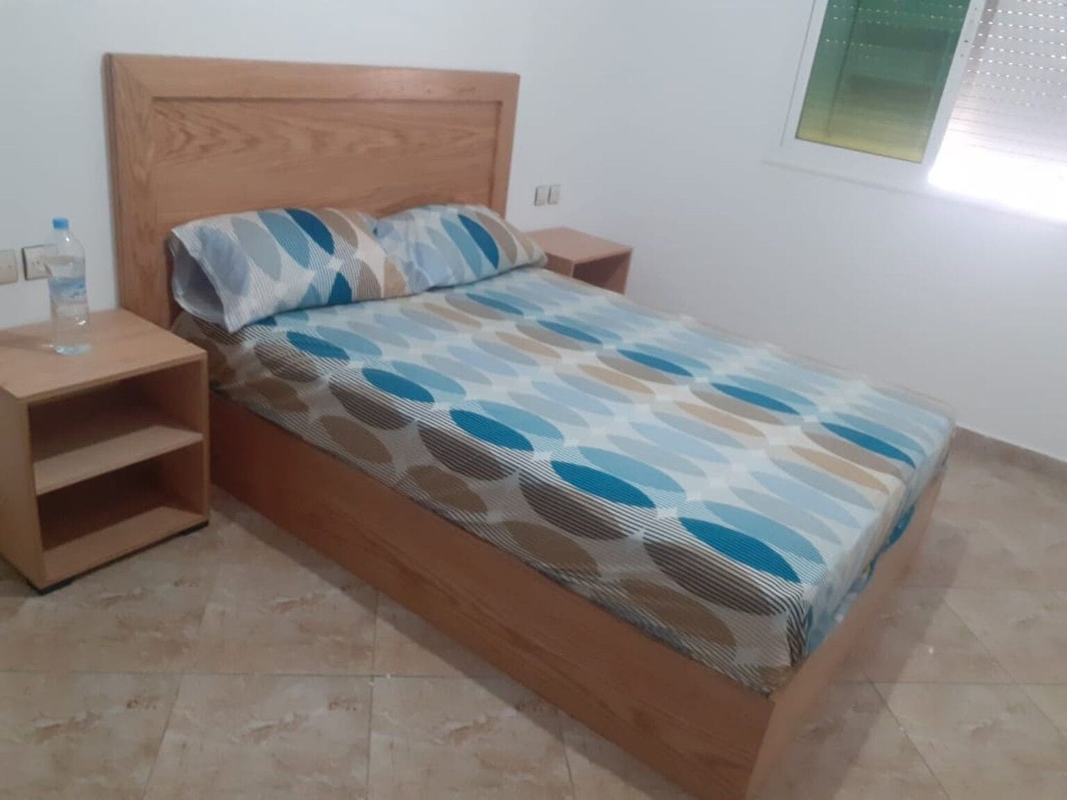Una cama con colcha estampada, cabecera de madera y mesita de noche con una botella de agua.
