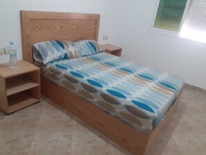 A bed with a patterned bedspread, wooden headboard, and a nightstand with a water bottle. - Immeuble Chaplin (Martil)