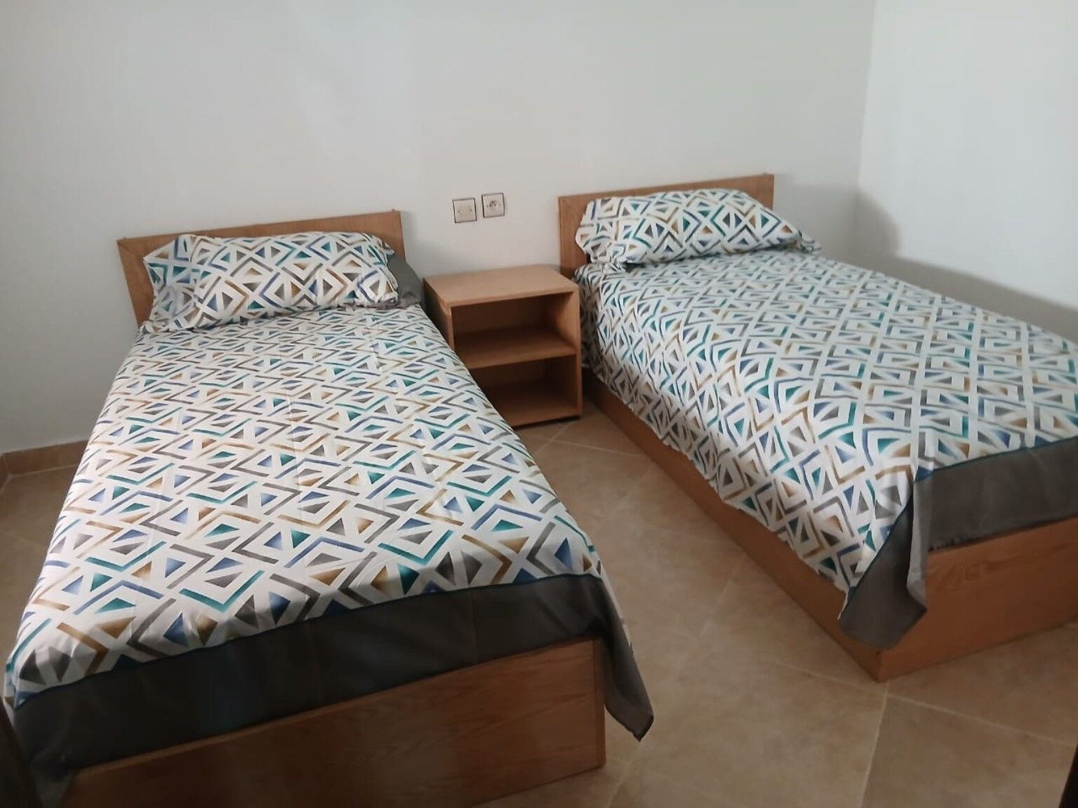 Two single beds with geometric patterned bedding, wooden headboards, and a small wooden nightstand between them.