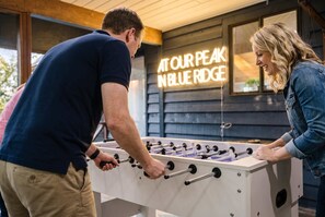A foosball table in a room with a neon sign that reads 'AT OUR PEAK BLUE RIDGE'. - Starry Skies: Sauna, S'mores & Pickleball Retreat (Blue Ridge)