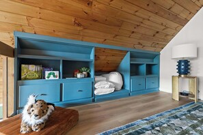 Een kamer met een schuine balkenlaag, een blauwe inbouwopbergruimte, een knuffel op een houten bankje en een kamerbreed tapijt met dessin.