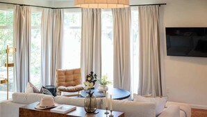 Ein Wohnzimmer mit einer Couch, einem Couchtisch und einem Sessel. Ein großes Fenster mit Vorhängen, eine Stehlampe und ein an der Wand befestigtes Fernsehgerät.