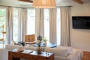 A living room with a sofa, a coffee table, and a chair. There is a large window with curtains, a floor lamp, and a wall-mounted TV. - Boutique 1BR Retreat, chilled & heated cowboy pool (Driftwood)