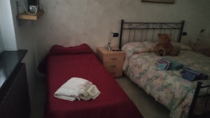 Quarto pequeno e simples com uma cama de solteiro, mesa de cabeceira, uma cadeira e um ursinho de pelúcia.