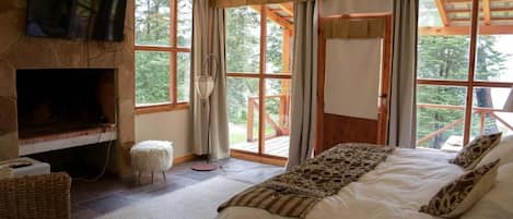 A bedroom with a stone fireplace, a bed with patterned bedding, and large windows offering a view of the outdoors.