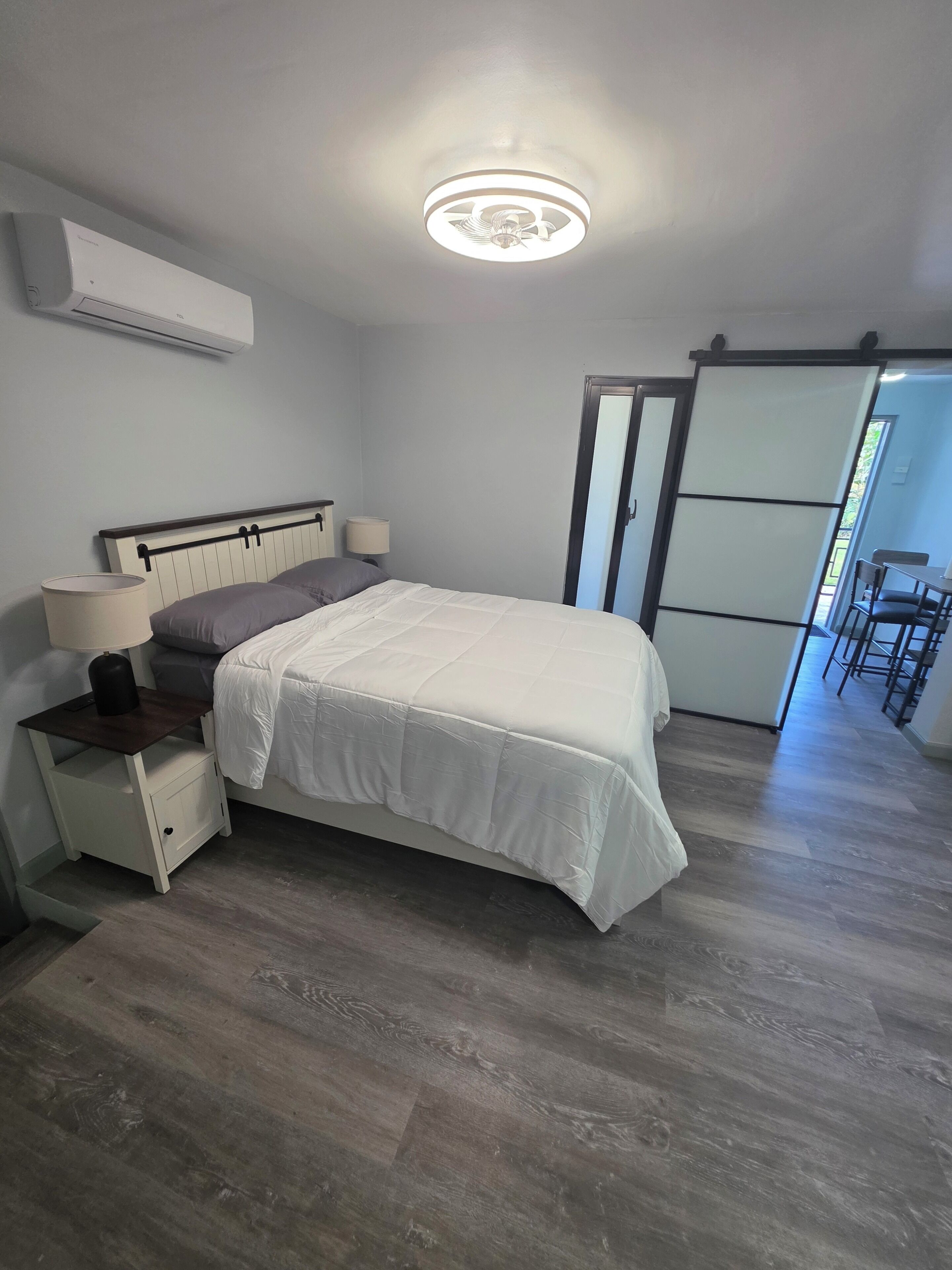 Un dormitorio con cama, mesitas de noche, lámpara, lámpara del techo y puerta corrediza que da a un comedor.