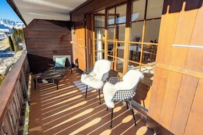 Eine Holzterrasse mit einer Couch und Stühlen, eine Glastür zum Innenbereich und Blick auf die Berge.